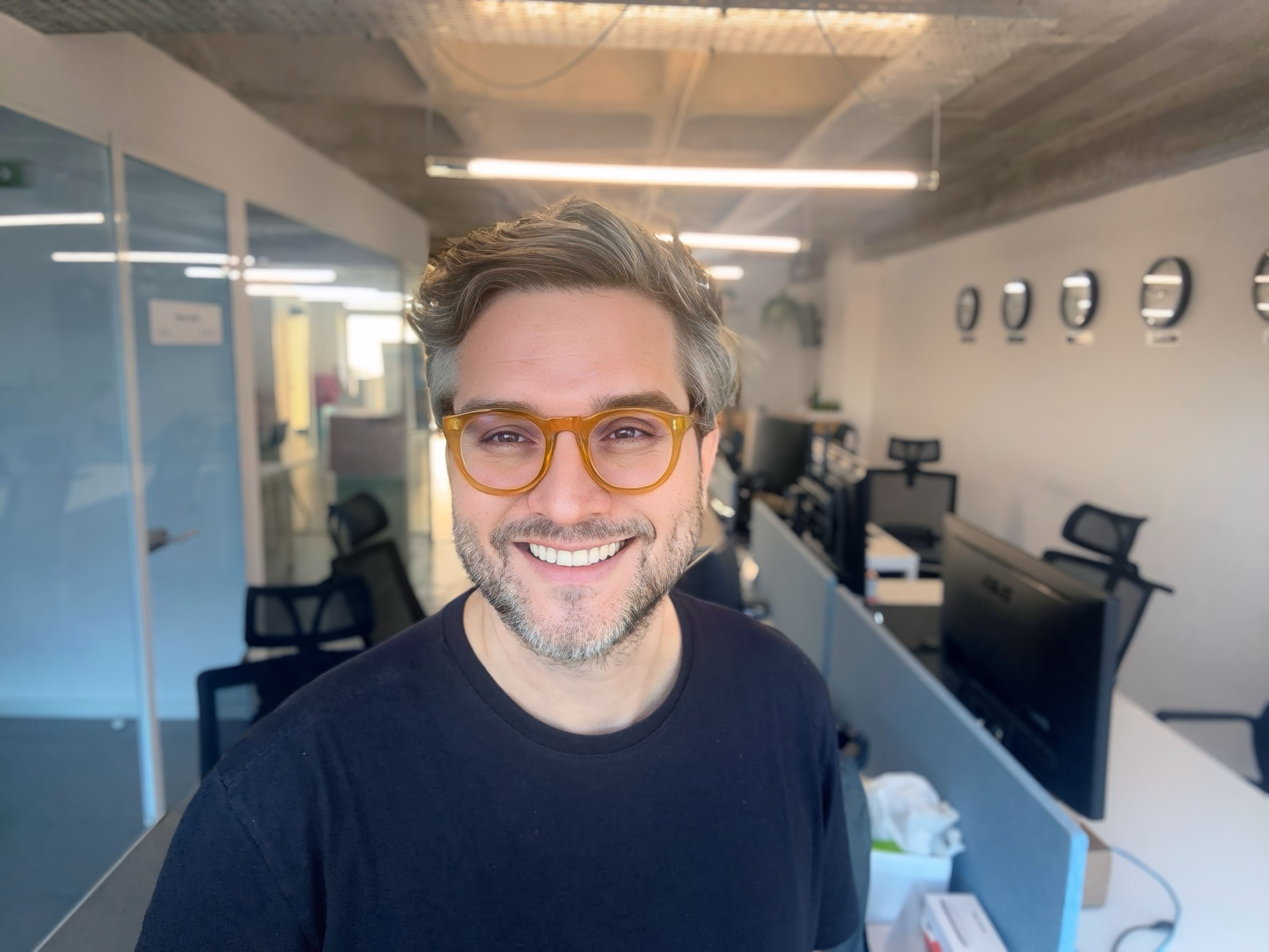 Ariel Alexandre sorrindo, de óculos âmbar, em primeiro plano em um escritório.